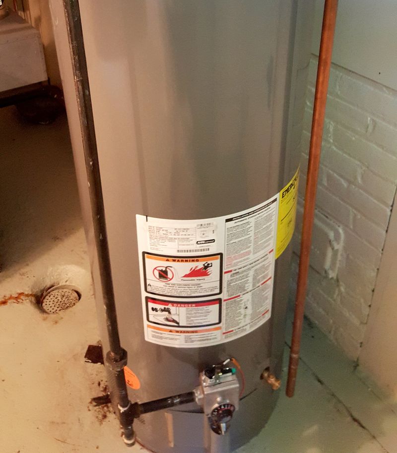 The lower half of a gas tank water heater in a basement, with the gas control valve near the bottom, the gas line, a copper relief-valve discharge pipe running down to the floor and a floor drain nearby